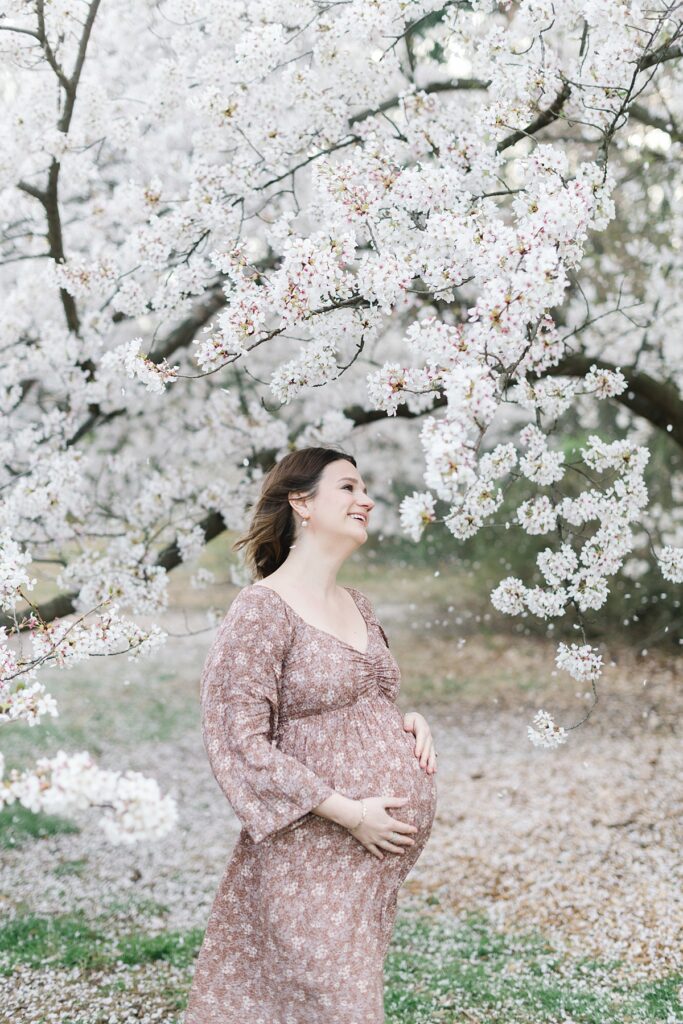 A pregnant woman in a muted pink and mauve floral print dress poses beneath a blooming tree at the National Arboretum for her maternity pictures
