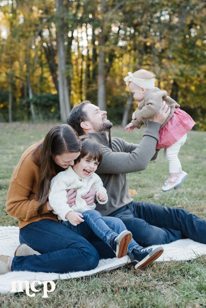 a family of four giggle, mom tickling the son, dad lifting the infant daughter up in the air as a candid image is taken during their fall family portrait session